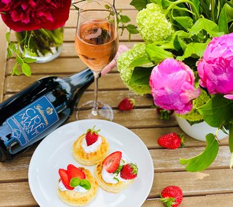 Ein Glas Rosé-Sekt steht neben einer Flasche Geldermann, umgeben von bunten Blumen und frischen Erdbeeren und Törtchen auf einem Holztisch.
