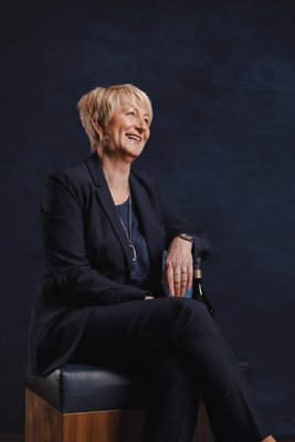 Die Frau trägt einen eleganten Anzug und zeigt eine freundliche Ausstrahlung, während sie auf einem Hocker sitzt.