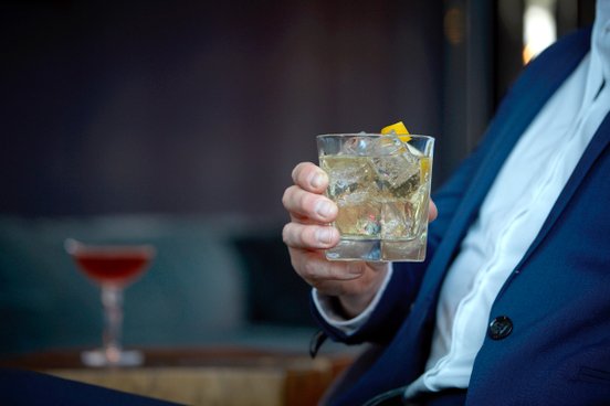 Ein Mann im blauen Anzug hält ein gefülltes Glas, das Eiswürfel und eine gelbliche Flüssigkeit hat