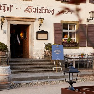 Vor dem Gasthaus 'Spielweg' steht ein Schild, das zu einem besonderen Abend einlädt, während ein Kellner die Tür öffnet.