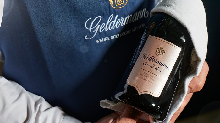 Eine Hand hält eine Flasche Geldermann Grand Rosé, umgeben von einem weißen Tuch, während ein blauer Hintergrund sichtbar ist.