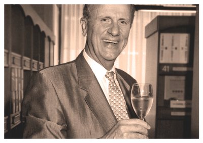 Ein älterer Mann in einem Anzug lächelt und hält ein Glas mit einem klaren Getränk in der Hand, umgeben von Bücherregalen.
