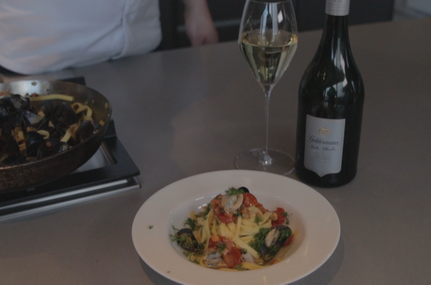 Ein Teller mit frischer Pasta, Muscheln und Tomaten, garniert mit Kräutern, steht auf einem Tisch neben einem Glas Weißwein.