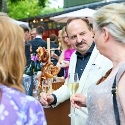 Johann Lafer genießt die Bayreuther Festspiel Cuvée 2025 im Gespräch mit anderen an einem Brezelstand