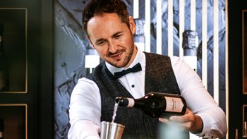 Eleganter Mann mit weißem Hemd und schwarzer Fliege schenkt auf Barthresen ein Glas ein