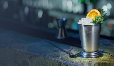 Der glänzende Becher mit Eis und Minze wird von einer Orangenscheibe dekoriert, während die Bar im Hintergrund verschwommen ist.