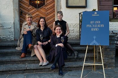 Vier Frauen sitzen entspannt auf einer Treppe vor einem historischen Gebäude, jede mit einem Glas Sekt in der Hand.