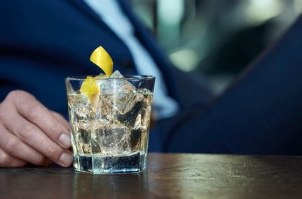 Eine Hand in einem Anzug hält ein Glas mit Eis und einem gelben Zitronenstreifen, während im Hintergrund unscharfe Farben zu sehen sind.