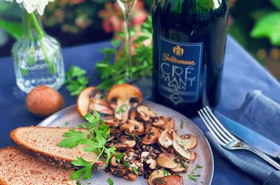 Ein Teller mit gebratenen Champignons, frischem Brot und einem Glas Crémant, umgeben von grünen Pflanzen im Hintergrund.