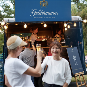 Eine fröhliche Frau hebt ein Glas Sekt an einem mobilen Getränkewagen mit dem Namen Geldermann, umgeben von Lichtern.