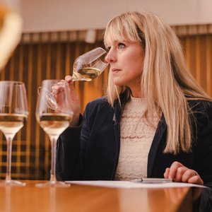 Eine Frau mit langen blonden Haaren genießt einen Schluck Weißwein aus einem Weinglas, während sie nachdenklich schaut.