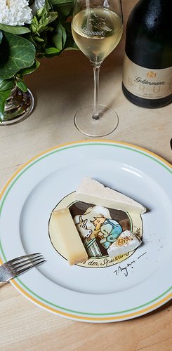 Auf einem weißen Teller liegen drei Käsestücke, während ein Glas Sekt und eine Blumenvase im Hintergrund sichtbar sind.