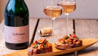 Zwei Gläser prickelnder Rosé stehen neben einer Flasche Geldermann und zwei appetitlichen Bruschetta auf einem Holzbrett.