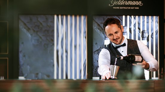 Der Mann im Anzug gießt mit einem Lächeln Sekt in einen Shaker, während eine stilvolle Bar im Hintergrund sichtbar ist.