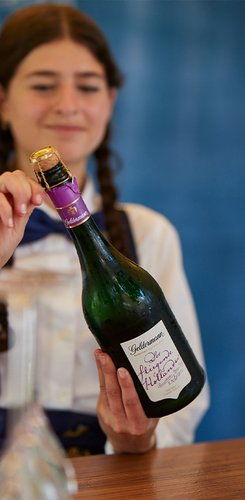 Eine junge Frau mit Zöpfen hält eine Sektflasche und lächelt, während sie den Korken öffnet.