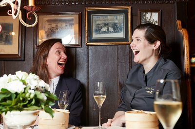 Zwei Frauen lachen herzlich am Tisch, umgeben von historischen Bildern und einer eleganten Tischdekoration mit Blumen.