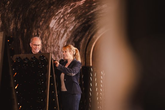 In einem stilvollen Weinkeller helfen sich ein Mann und eine Frau, während sie an einem Regal mit Weinflaschen arbeiten und lachen.