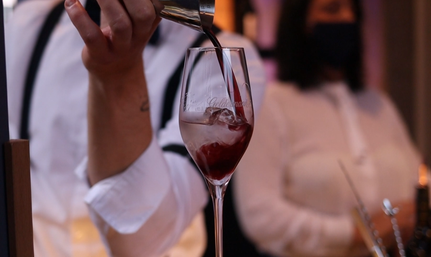 Ein Glas mit Eis wird mit einem roten Getränk gefüllt, während eine Person im Hintergrund in einem weißen Oberteil und einer Maske zu sehen ist.