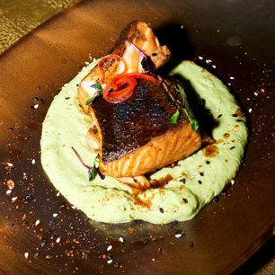 Ein perfekt gegrilltes Stück Lachs liegt auf einer cremigen, grünen Sauce, garniert mit frischen Kräutern und roten Chilischoten.