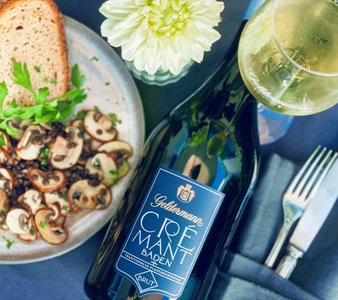 Ein eleganter Tisch mit einem Glas Crémant, einer Flasche und einem Teller mit Pilzen und frischem Brot, umgeben von Kräutern.