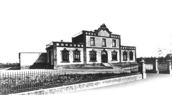 Ein historisches Gebäude mit einer markanten Fassade und mehreren Fenstern, umgeben von einem schmiedeeisernen Zaun.