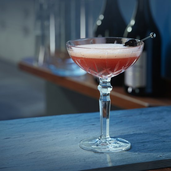 Ein eleganter Cocktail in einem Kristallglas steht auf einer blauen Theke, im Hintergrund sind Flaschen sichtbar.