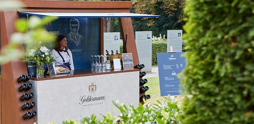 Eine Frau steht an einem stilvollen Verkaufsstand mit Weinflaschen und Blumen, umgeben von grünen Pflanzen im Park.