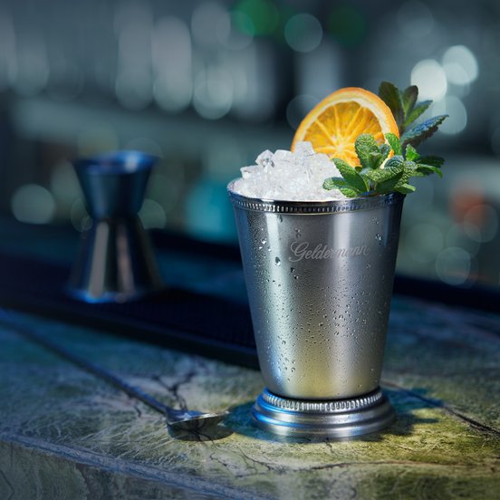 Der glänzende Becher, beschlagen mit Wassertropfen, enthält Eis und wird von einer Orangenscheibe und Minze dekoriert.