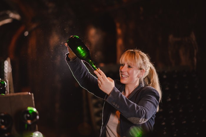 Eine blonde Frau in einem Weinkeller hebt eine Flasche, während sie von weiteren Flaschen umgeben ist und lächelt.