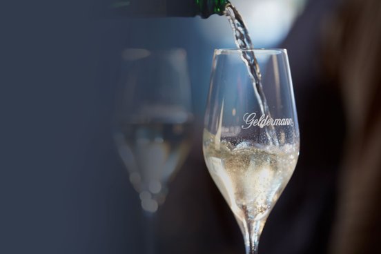 Der Schaum eines prickelnden Getränks steigt auf, während es in ein elegantes Glas mit dem Namen 'Geldermann' gegossen wird.