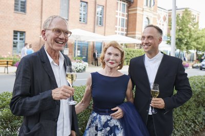 Ein älterer Mann, eine Frau mit roten Haaren und ein jüngerer Mann lächeln fröhlich, während sie an einem festlichen Anlass teilnehmen.