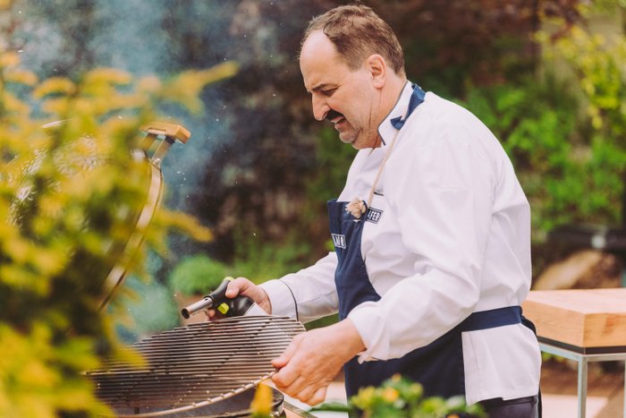 Ein Koch in einem weißen Hemd und blauen Schürze zündet mit einem Brenner den Grill im Garten an, um das Essen vorzubereiten.