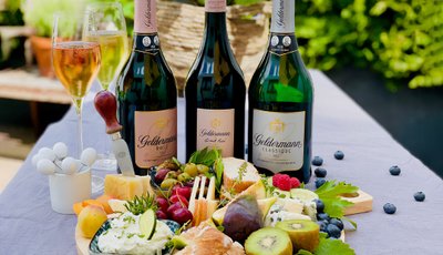 Ein gedeckter Tisch im Freien zeigt eine Auswahl an Käse, frischen Früchten und drei Flaschen Geldermann Sekt.