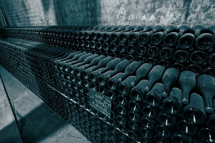 Eine große Ansammlung von Weinflaschen liegt ordentlich gestapelt in einem dunklen Keller, umgeben von rauen Wänden.