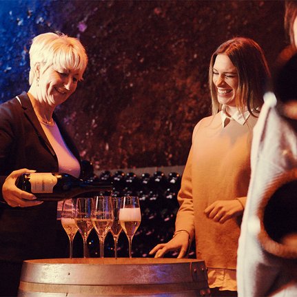 Eine blonde Frau schenkt Sekt in Gläser, während eine andere Frau lächelt und die Atmosphäre in einem Weinkeller genießt.