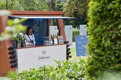 Eine Frau steht an einem stilvollen Verkaufsstand mit Weinflaschen und Blumen, umgeben von grünen Pflanzen im Park.
