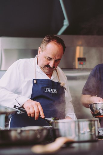 Der Koch, gekleidet in eine weiße Jacke und eine blaue Schürze, konzentriert sich auf das Kochen, während Dampf aus dem Topf aufsteigt.