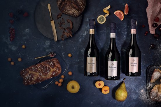 Ein stilvolles Arrangement mit Sektflaschen, frischen Früchten und einem goldenen Besteckstück auf einem dunklen Hintergrund.