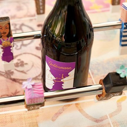Eine Flasche Geldermann Sekt steht im Mittelpunkt, umgeben von bunten Figuren auf einem Tischkicker mit floralem Muster.