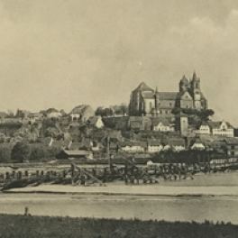 Eine malerische Landschaft mit einer Stadt auf einem Hügel, umgeben von Wasser und einer Holzbrücke im Vordergrund.