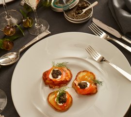 Drei kunstvoll angerichtete Häppchen auf einem weißen Teller, garniert mit Dill und schwarzem Kaviar, umgeben von eleganten Gläsern.