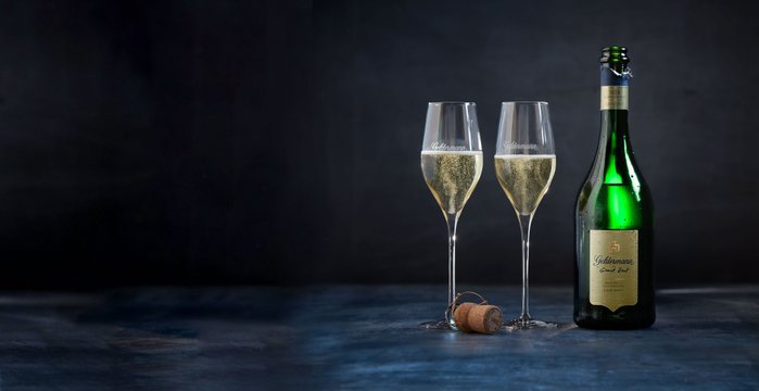 Zwei elegante Sektgläser mit sprudelndem Inhalt stehen neben einer grünen Sektflasche auf einem dunklen Hintergrund.