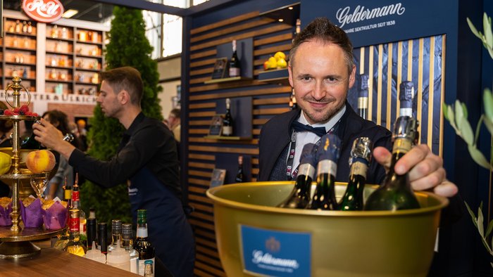 Ein lächelnder Mann in einem Anzug hält eine goldene Schüssel mit Sektflaschen, während im Hintergrund eine Bar zu sehen ist.