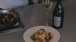 Ein Teller mit frischer Pasta, Muscheln und Tomaten, garniert mit Kräutern, steht auf einem Tisch neben einem Glas Weißwein.