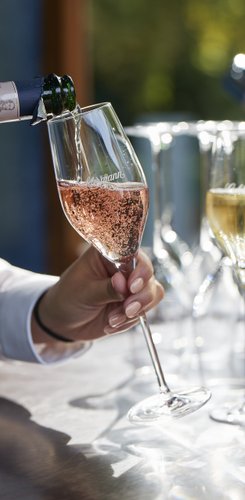 Ein Glas mit sprudelndem Rosé wird von einer Hand eingeschenkt, während weitere Gläser im Hintergrund stehen.