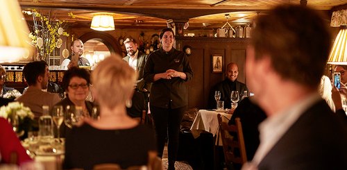 Eine lächelnde Köchin spricht zu den Gästen in einem gemütlichen Restaurant mit warmem Licht und rustikalem Ambiente.