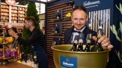 Ein lächelnder Mann in einem Anzug hält eine goldene Schüssel mit Sektflaschen, während im Hintergrund eine Bar zu sehen ist.
