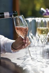 Ein Glas mit sprudelndem Rosé wird von einer Hand eingeschenkt, während weitere Gläser im Hintergrund stehen.
