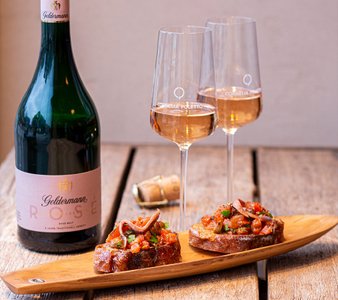 Eine Flasche Geldermann Rosé Brut steht neben zwei Gläsern Sekt und einer Holzplatte mit belegtem Brot auf einem Tisch.