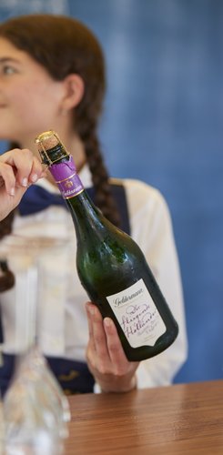 Die Hand eines Mädchens greift nach dem Korken einer Sektflasche, während Gläser auf einem Holztisch bereitstehen.